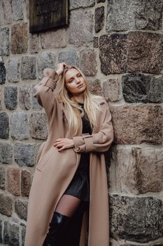 Elegant portrait of a fashionable blonde woman in a beige woolen coat, standing against a stone wall in an urban setting.