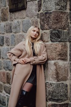 Stylish blonde woman in a beige coat poses elegantly against a rustic stone wall.