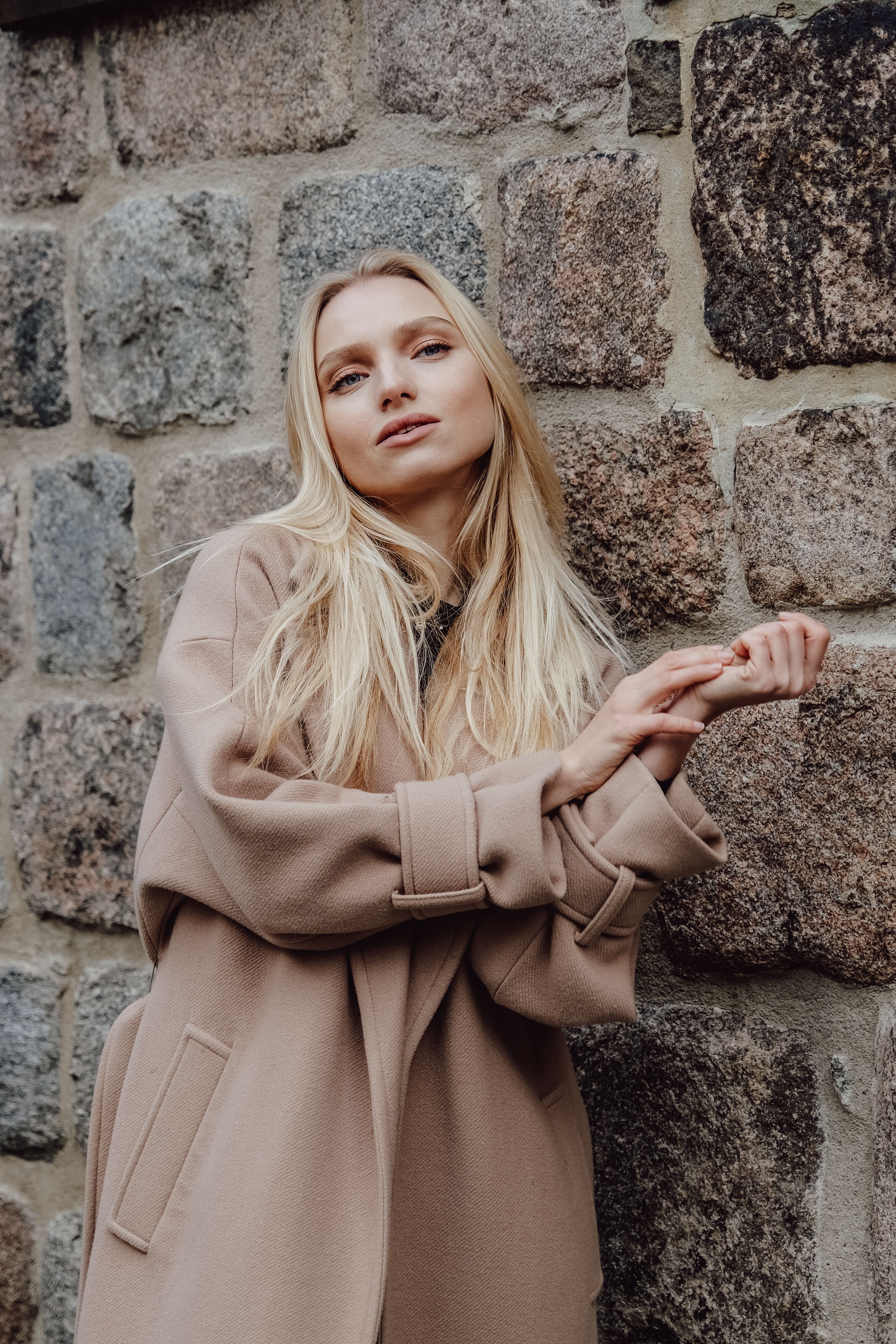 Fashion portrait of a woman in a beige coat posing stylishly against a rustic stone wall.