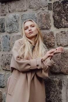 Fashion portrait of a woman in a beige coat posing stylishly against a rustic stone wall.