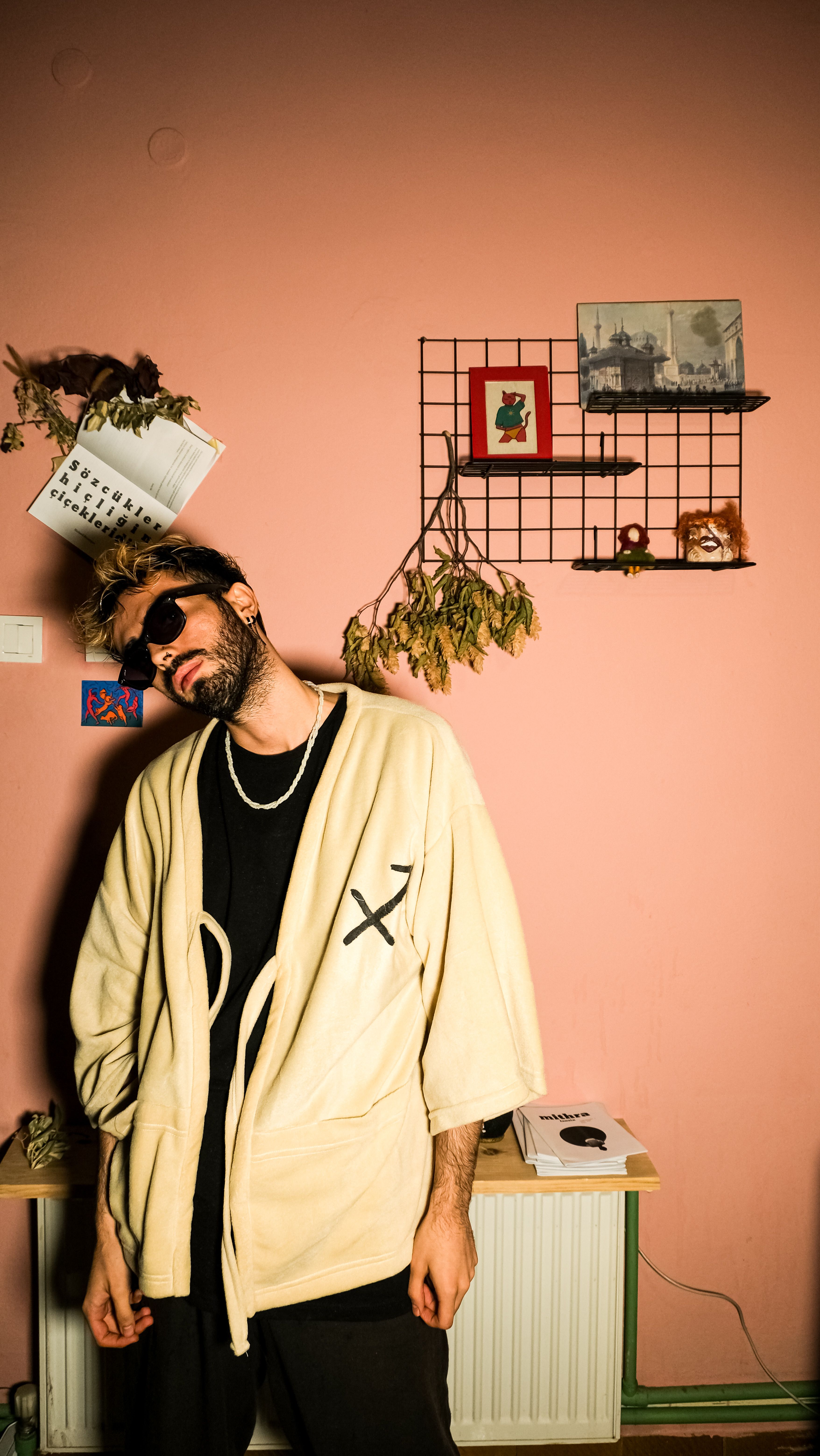 Stylish man in sunglasses and cardigan poses against a pink wall, showcasing modern fashion.