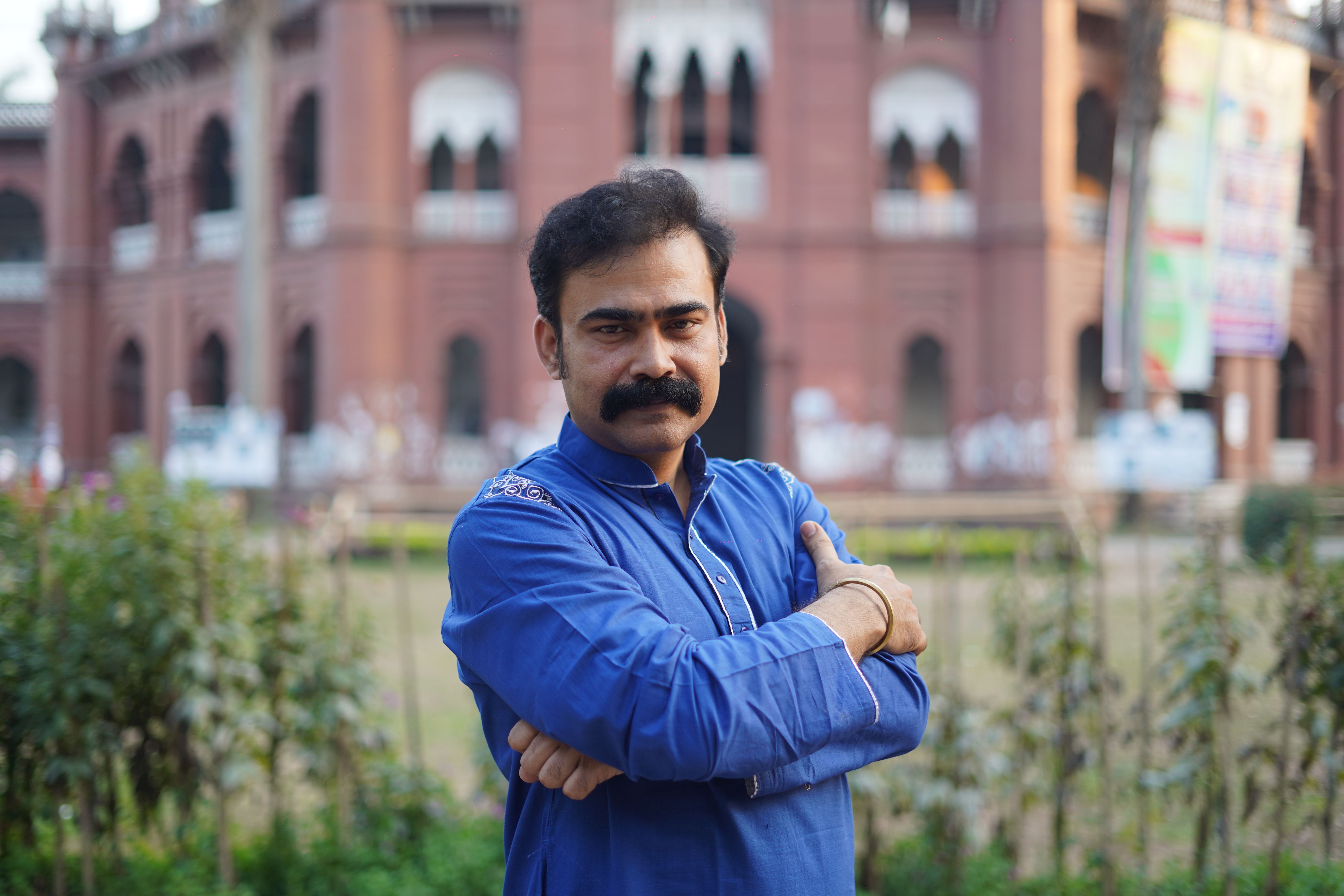 Portrait of a man in a blue shirt standing confidently outdoors with a historic red brick building in the background.