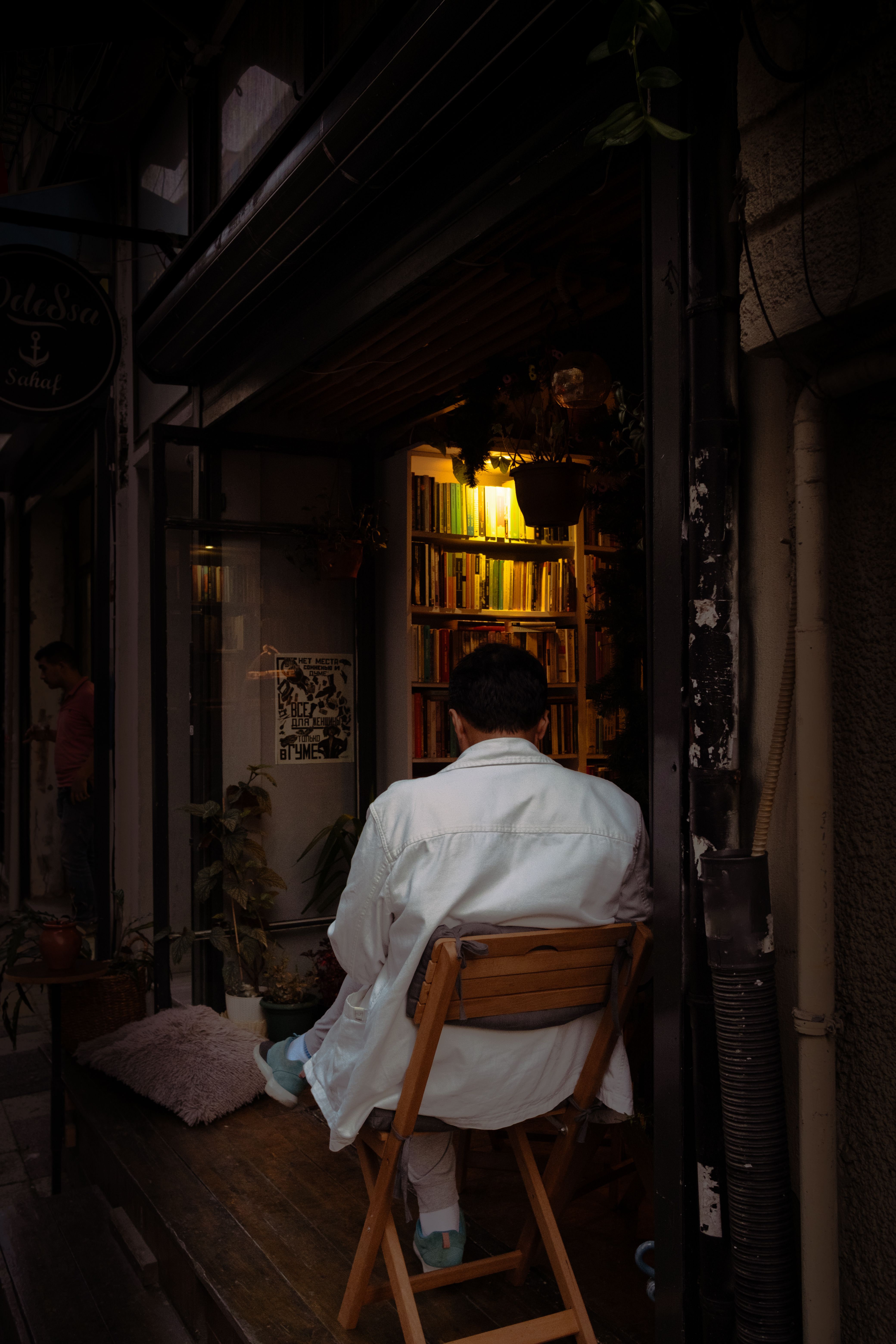 A man in a white shirt sits on a chair inside a warm, inviting bookstore at night.