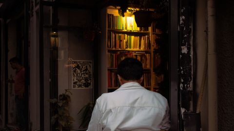 A man in a white shirt sits on a chair inside a warm, inviting bookstore at night.
