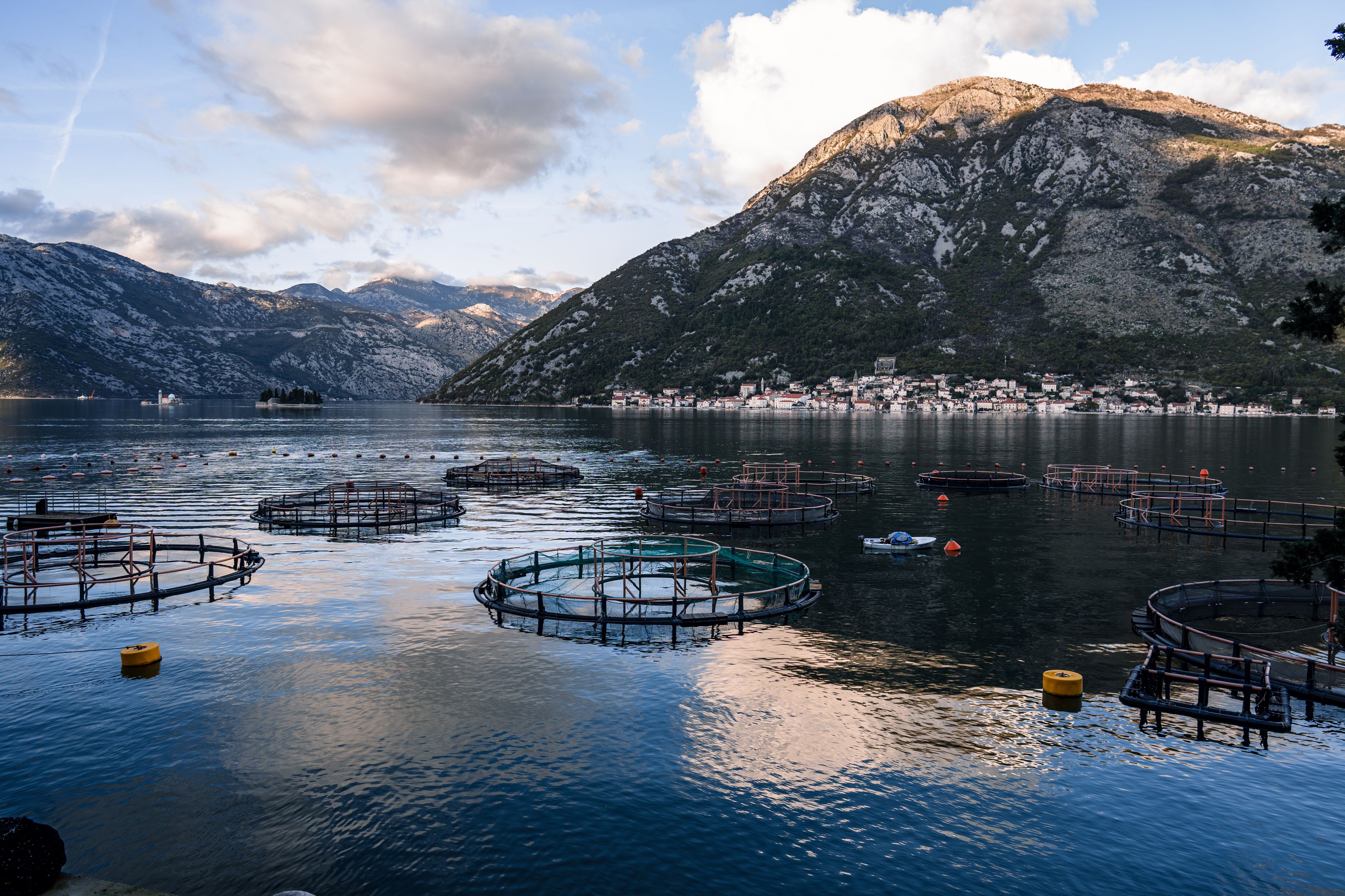Serene mountain bay with fish farms under a partly cloudy sky.