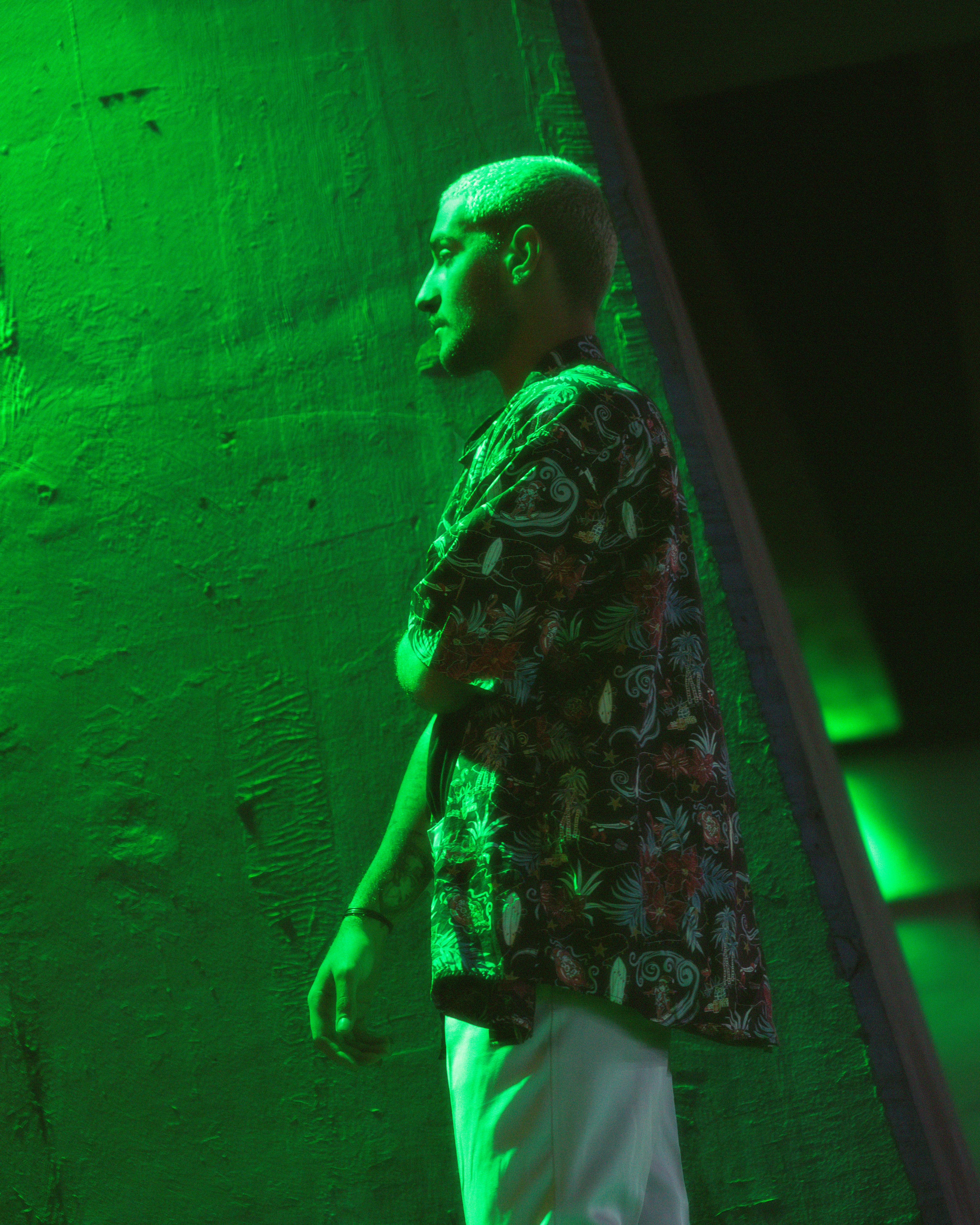 Fashionable model stands against a green-lit urban wall, showcasing modern style.