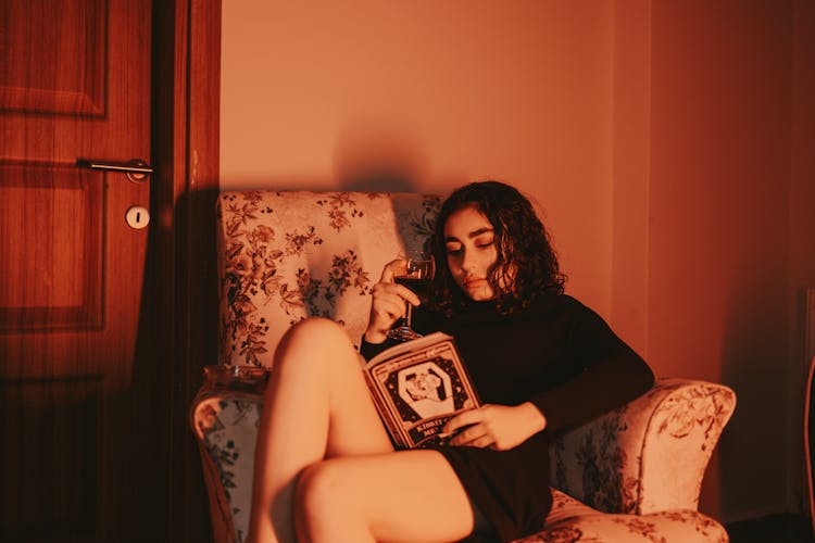 Brunette Woman In Black Mini Dress Reading A Book With A Glass Of Wine