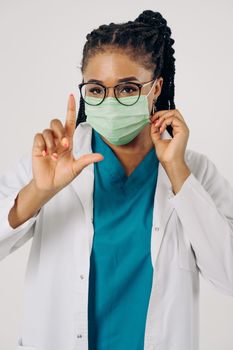 Female doctor in scrubs and surgical mask gesturing a sign in a studio setting.