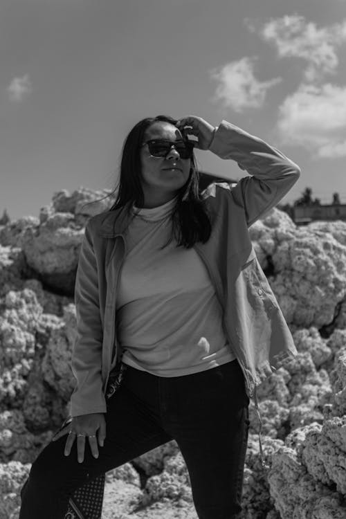 Black and White Photo of a Woman in Sunglasses Standing on Rocks 