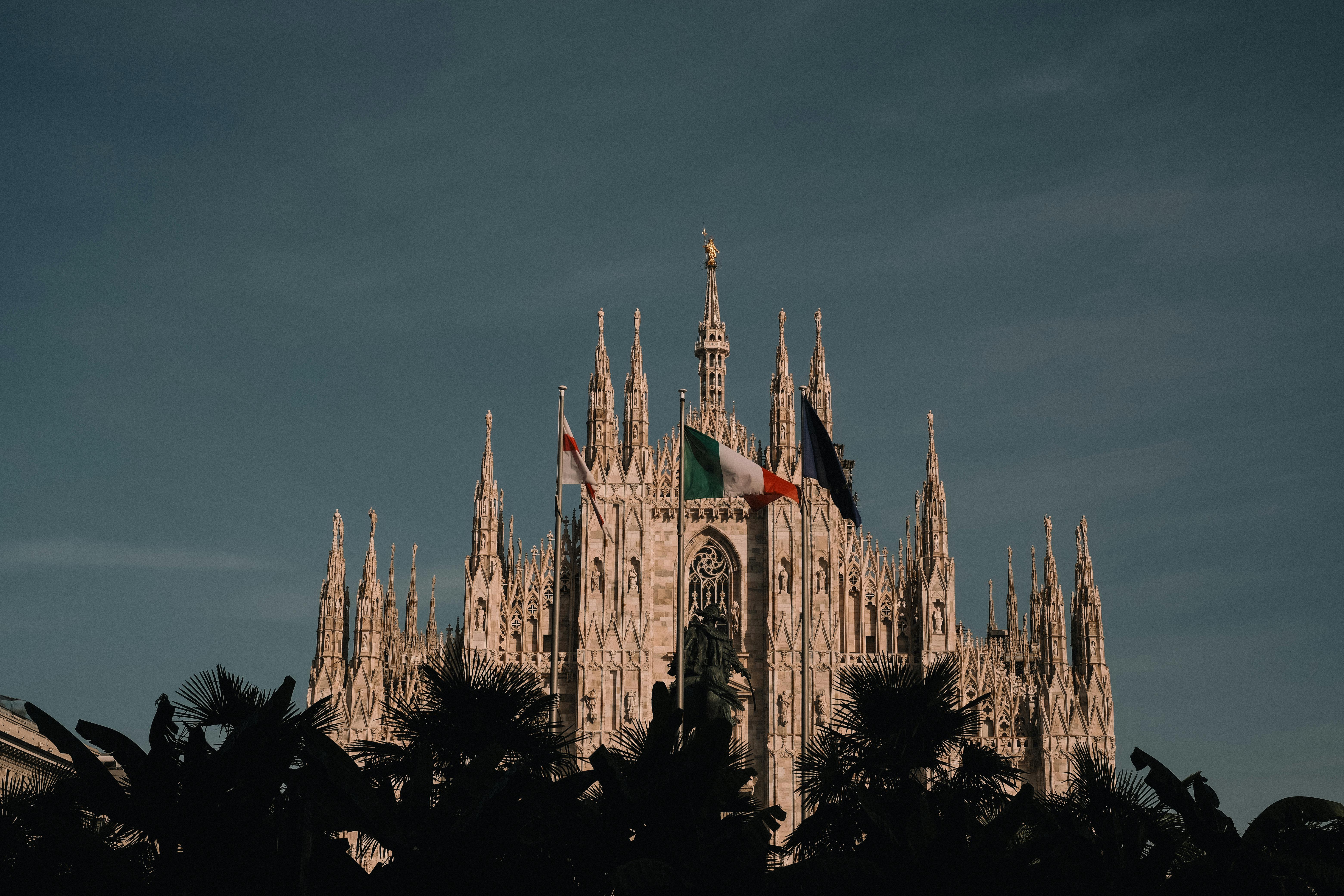 Gothic architecture of Milan Cathedral against a dark sky adorned by Italian flags.