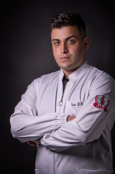Portrait of a serious man in a white jacket with arms crossed, set against a gray studio background.