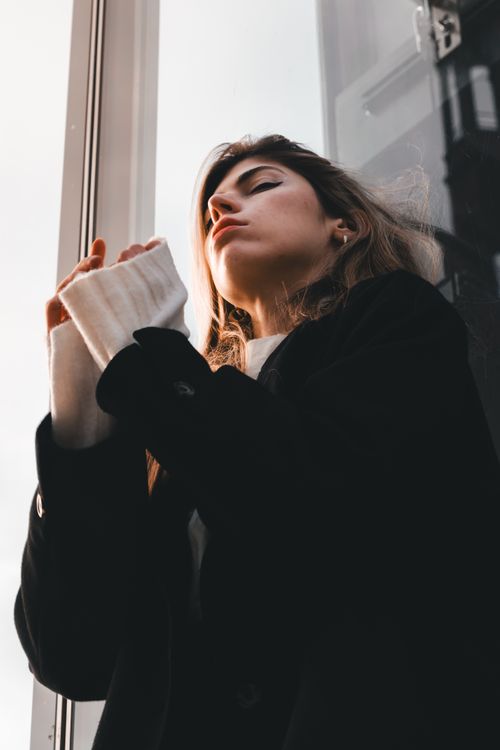 Free Portrait of a young woman in a black coat standing by a large window. Stock Photo