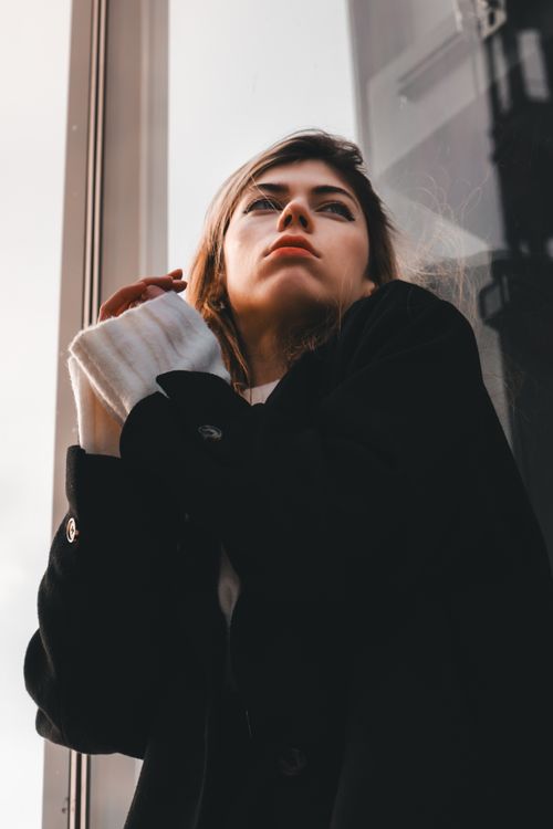 Free A thoughtful woman in a black coat gazing up beside a window, exuding fashion and elegance. Stock Photo