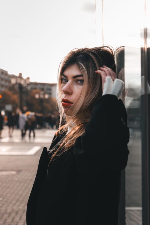 Free Woman with blonde hair in a black coat poses by a city building, reflecting urban elegance. Stock Photo