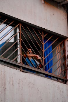 在城市环境中，一名男子从装有防护栏的窗户向外望去，捕捉着日常生活的瞬间
