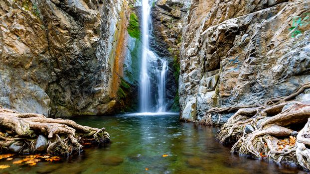 Majestic waterfall cascading down rocky cliffs into a serene pool with exposed roots and lush greenery.