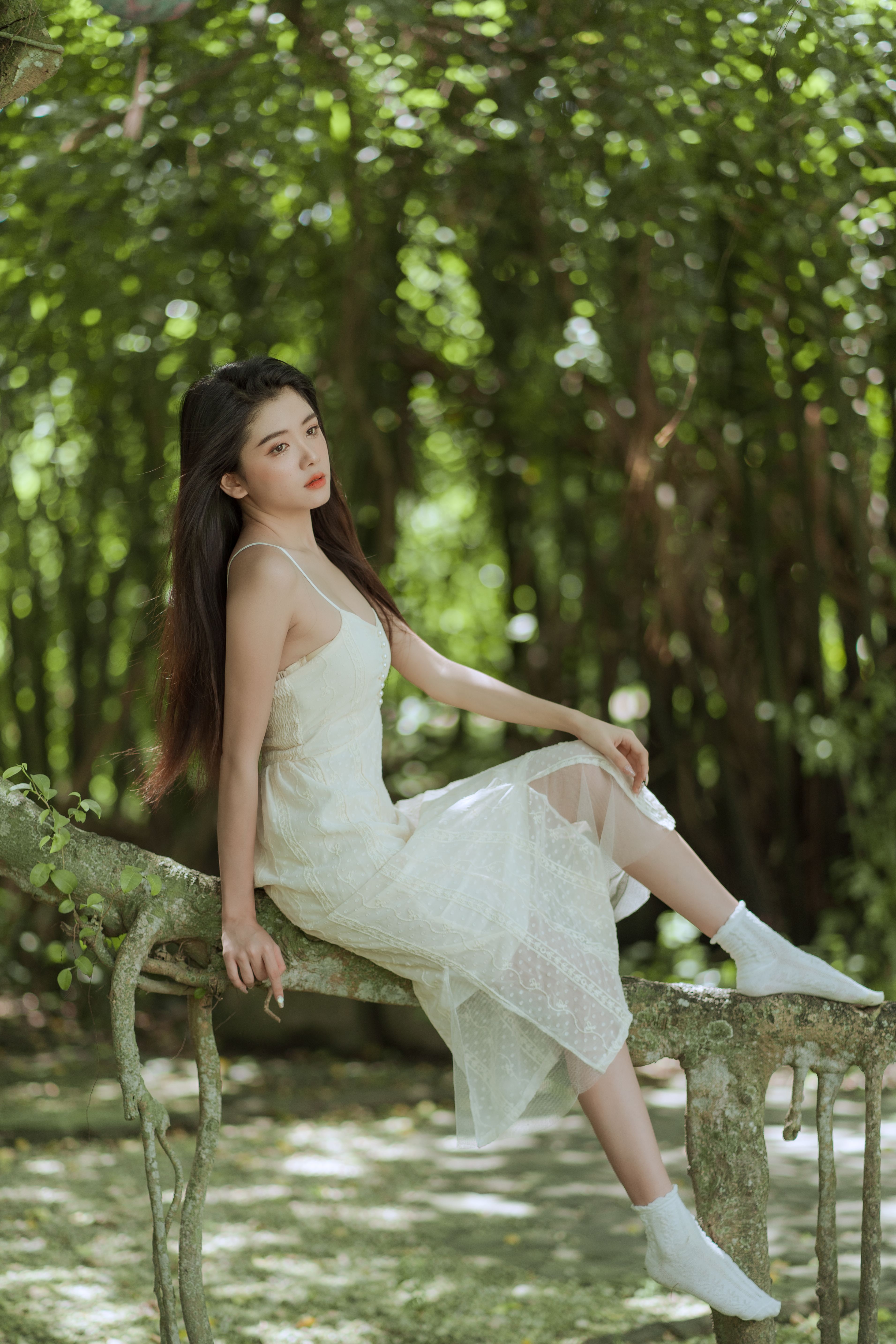 Beautiful woman in white, seated gracefully on a branch in a serene, sunlit park setting.