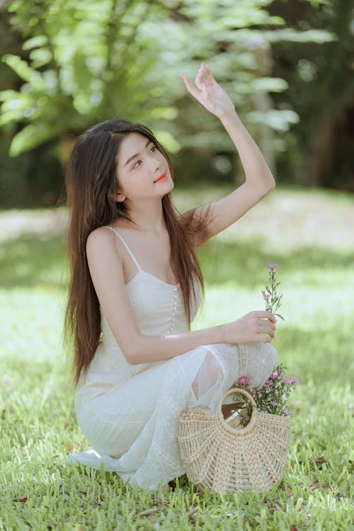 Model in a White Spaghetti Strap Summer Dress with Wicker Handbag on a Grass in the Park