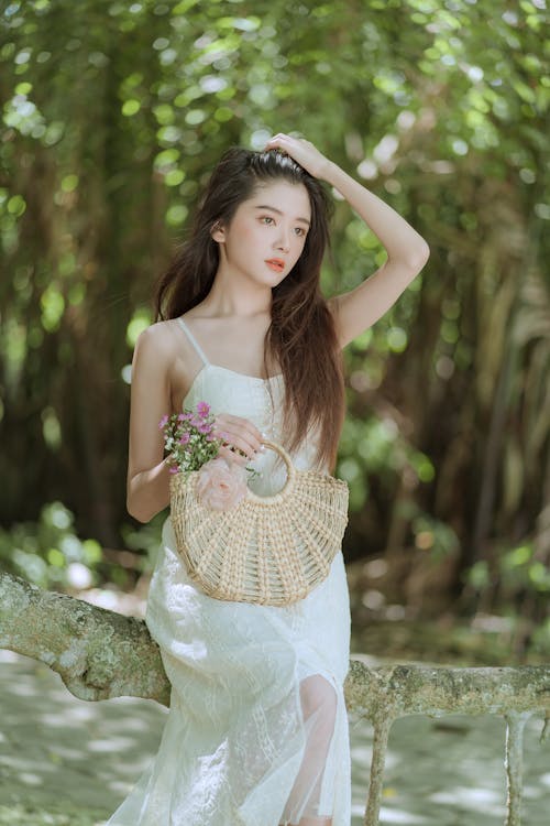 Model in a White Spaghetti Strap Summer Dress with a Wicker Handbag Sitting on a Branch in the Park