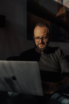 Bearded man with eyeglasses working on a laptop in a dimly lit room, focusing intently.
