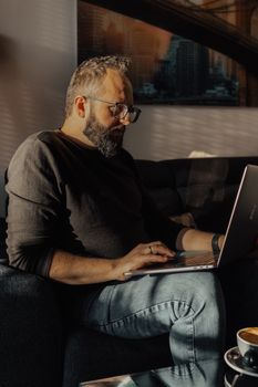 Man with beard and glasses using a laptop on sofa, sunlight streaming indoors.
