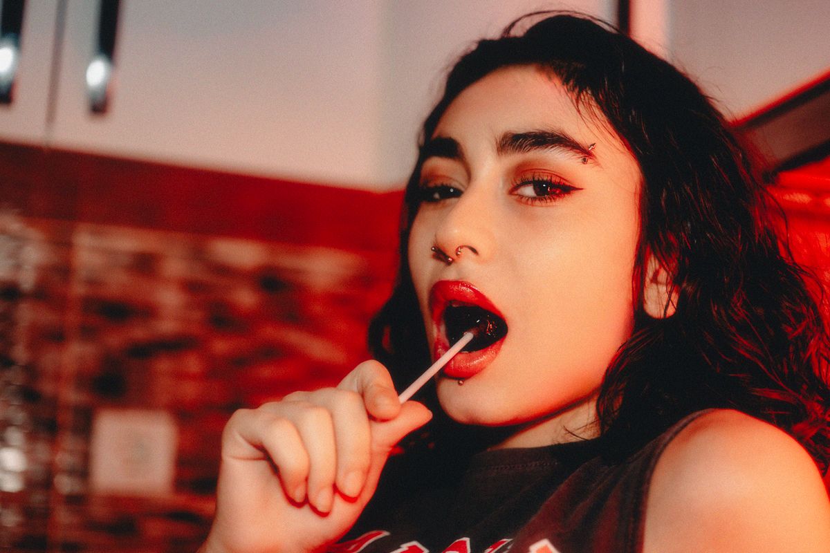 Close-up portrait of a fashionable woman with piercings and red lipstick enjoying a lollipop indoors.