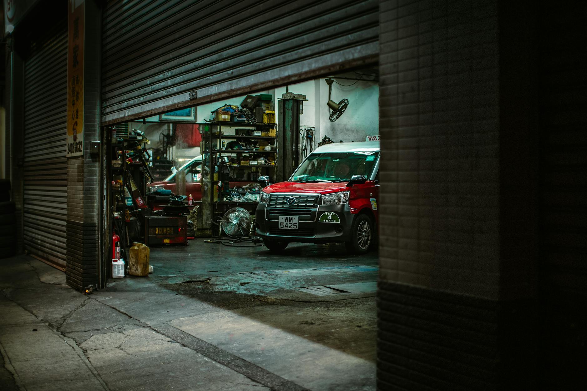 Red car inside a Hong Kong auto repair shop with open roller shutter, showcasing tools and equipment.