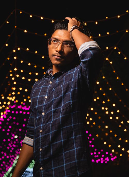 Free A young man in a checkered shirt poses under glowing string lights in an outdoor evening setting. Stock Photo