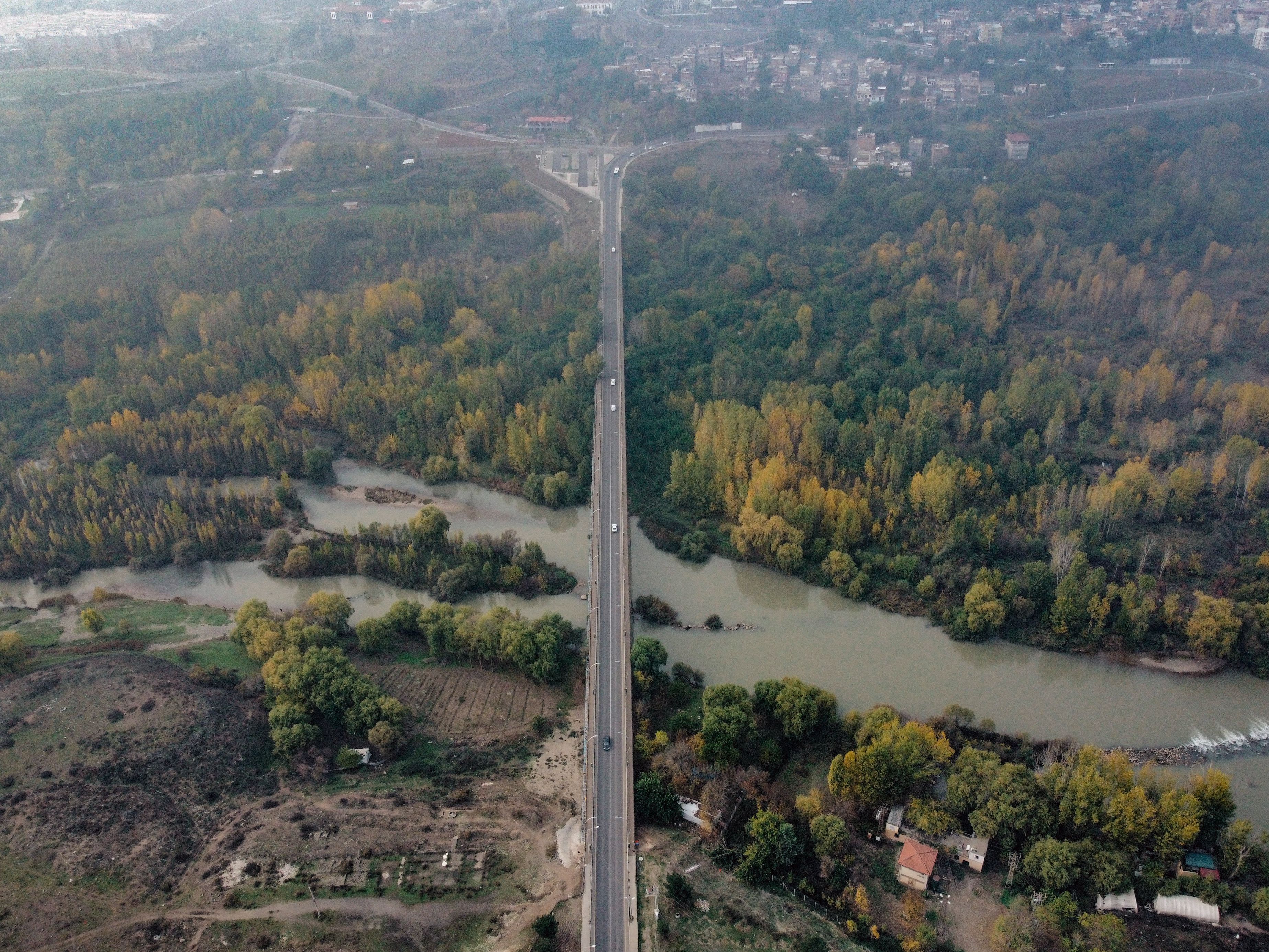 Drone shot capturing a bridge spanning a river amidst lush green forest and suburban area.