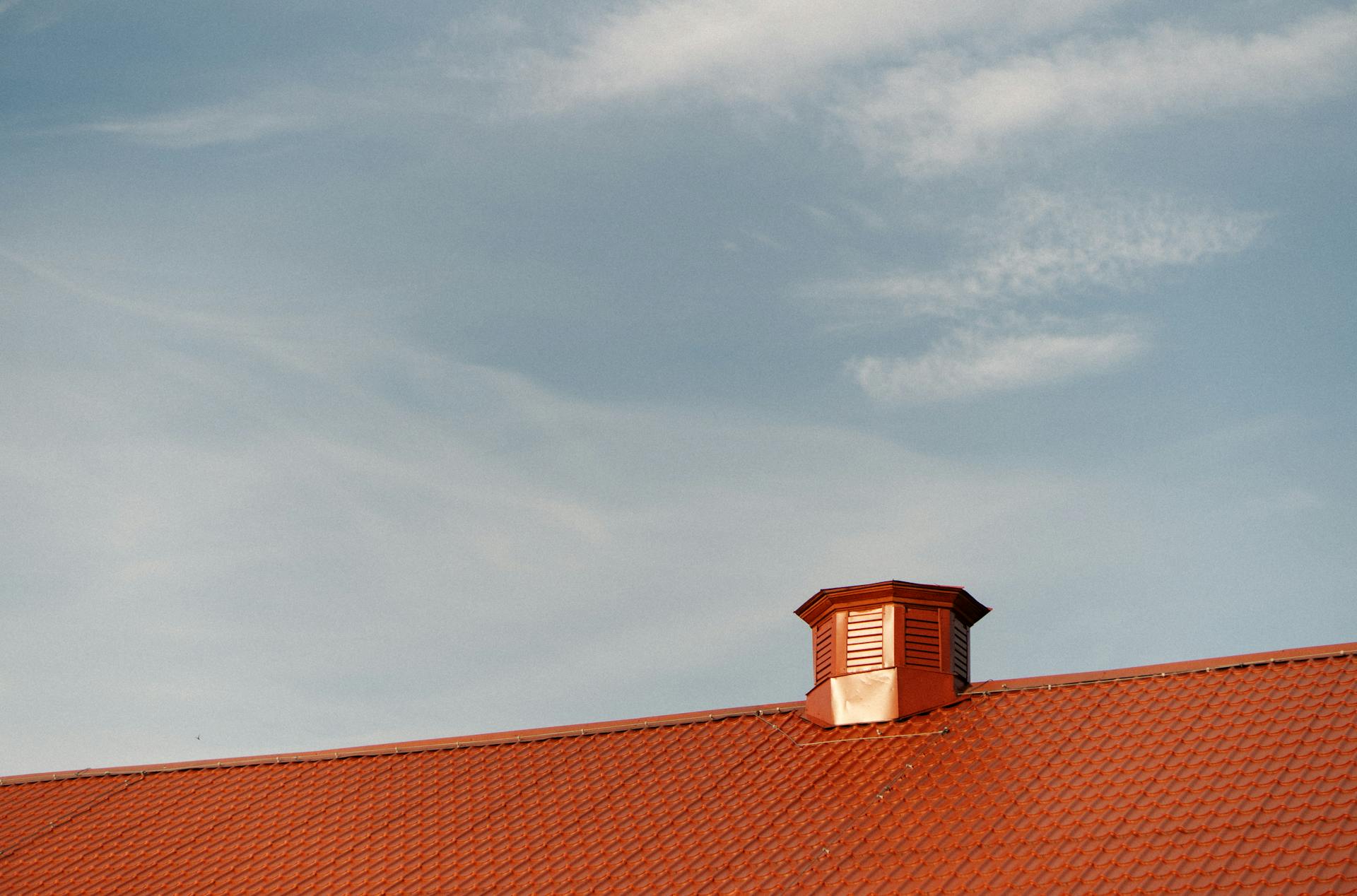 A serene view of a red tin rooftop with a vent under a clear blue sky.