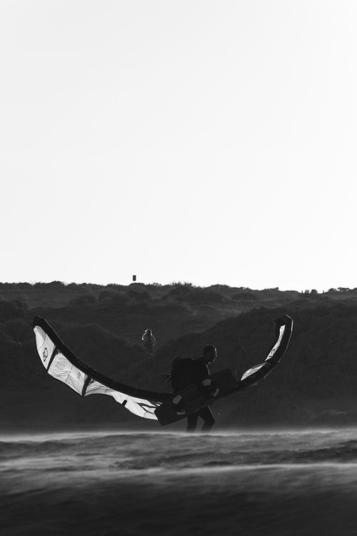 Free A striking black and white silhouette of a kitesurfer carrying equipment along a windy seaside at dusk. Stock Photo