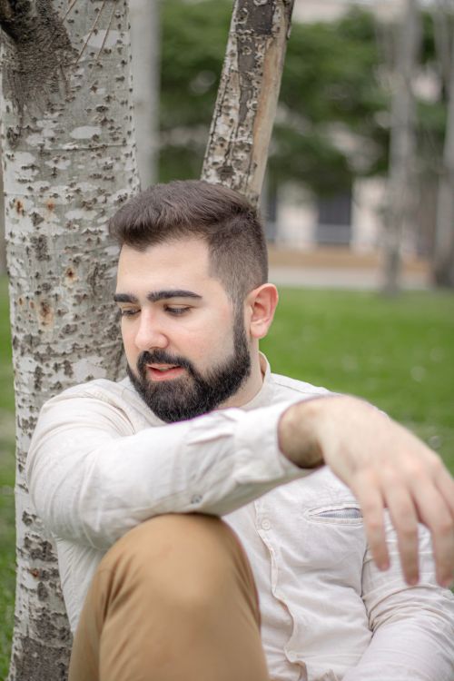 Free A young bearded man sitting and relaxing against a tree in a park setting. Stock Photo