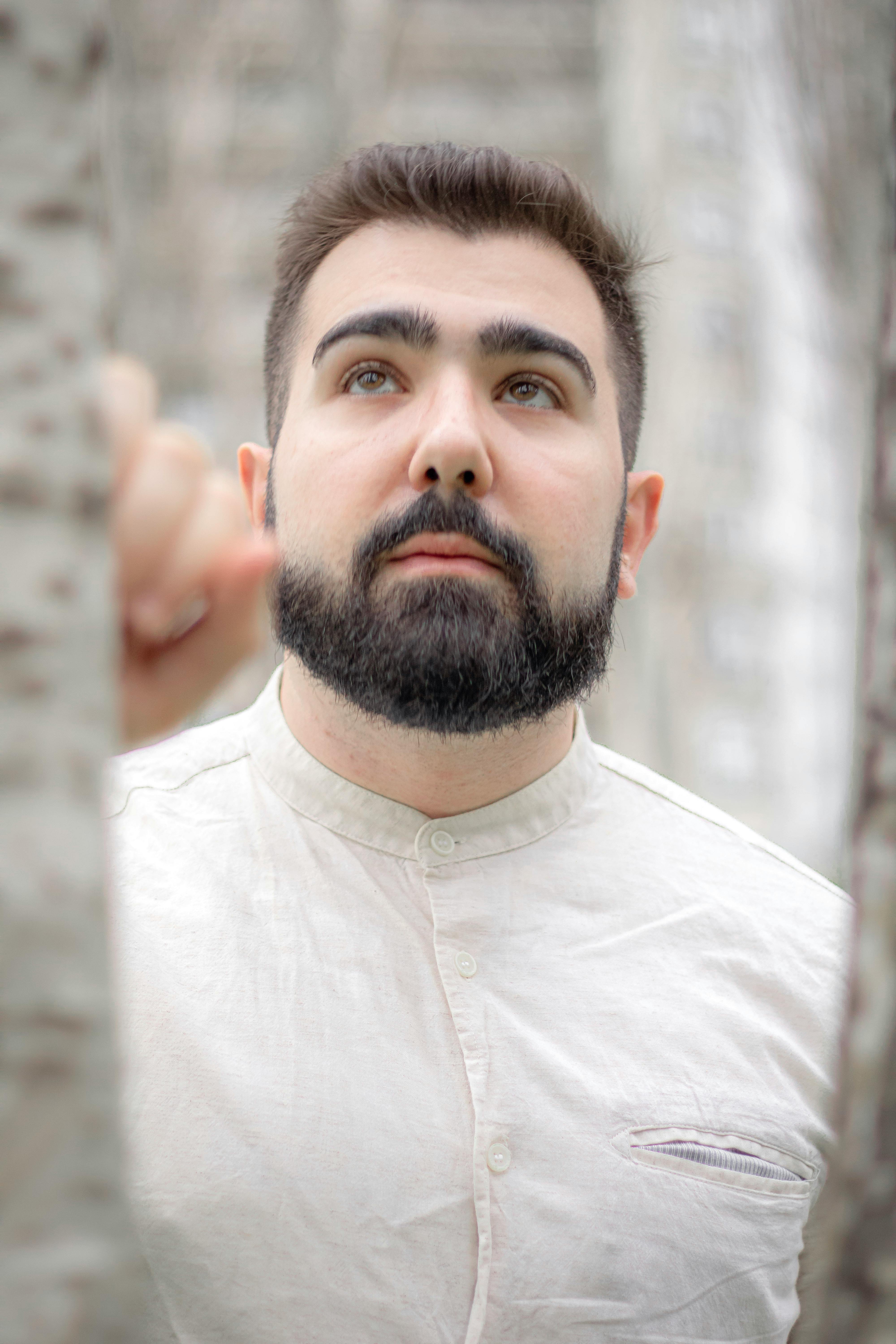 Close-up portrait of a bearded man in a white shirt, looking away pensively in an outdoor setting.