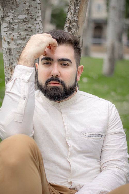 Free A thoughtful man with a beard sitting outdoors against a tree. Stock Photo