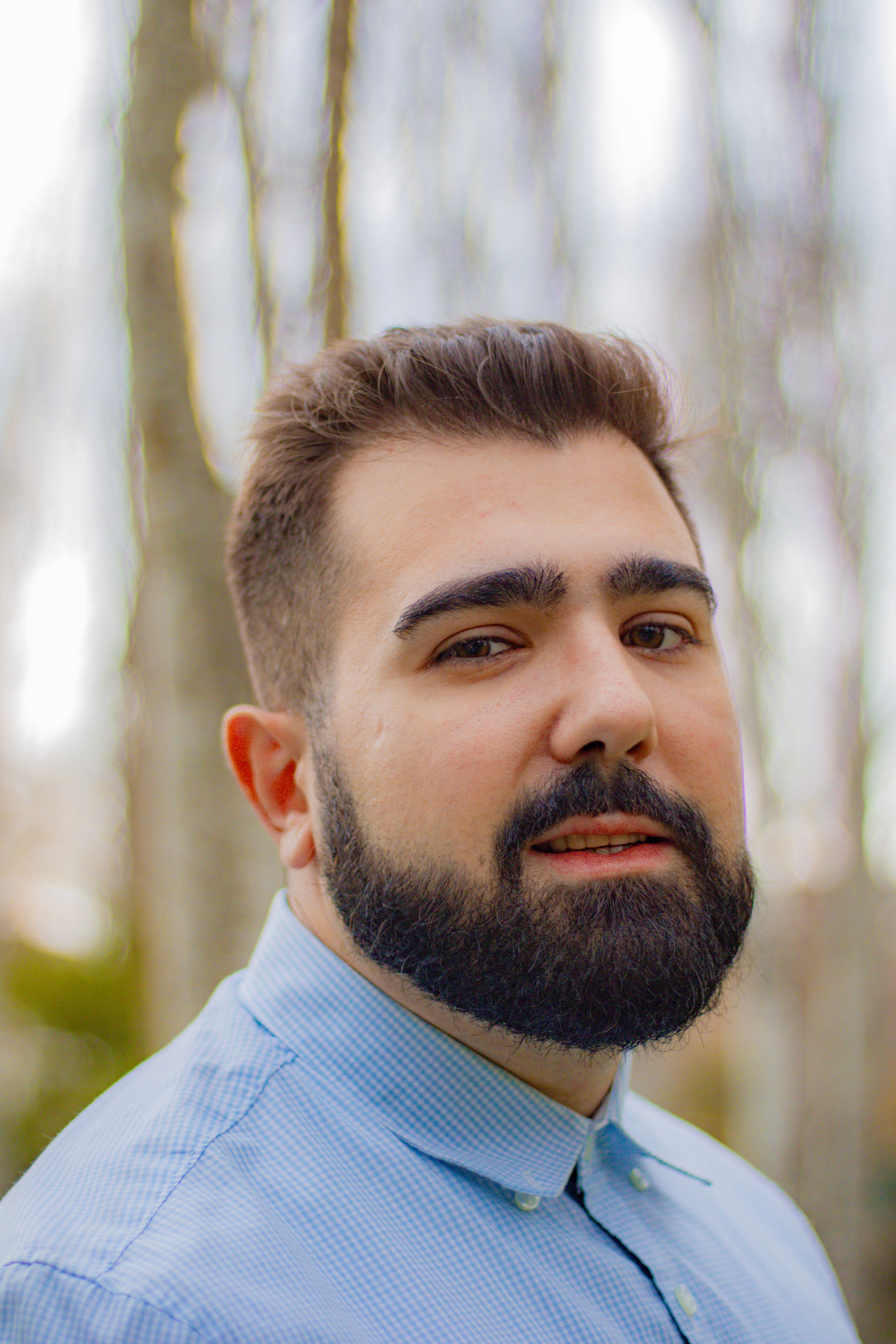 Close-up portrait of a bearded man with a blue shirt in a natural setting.