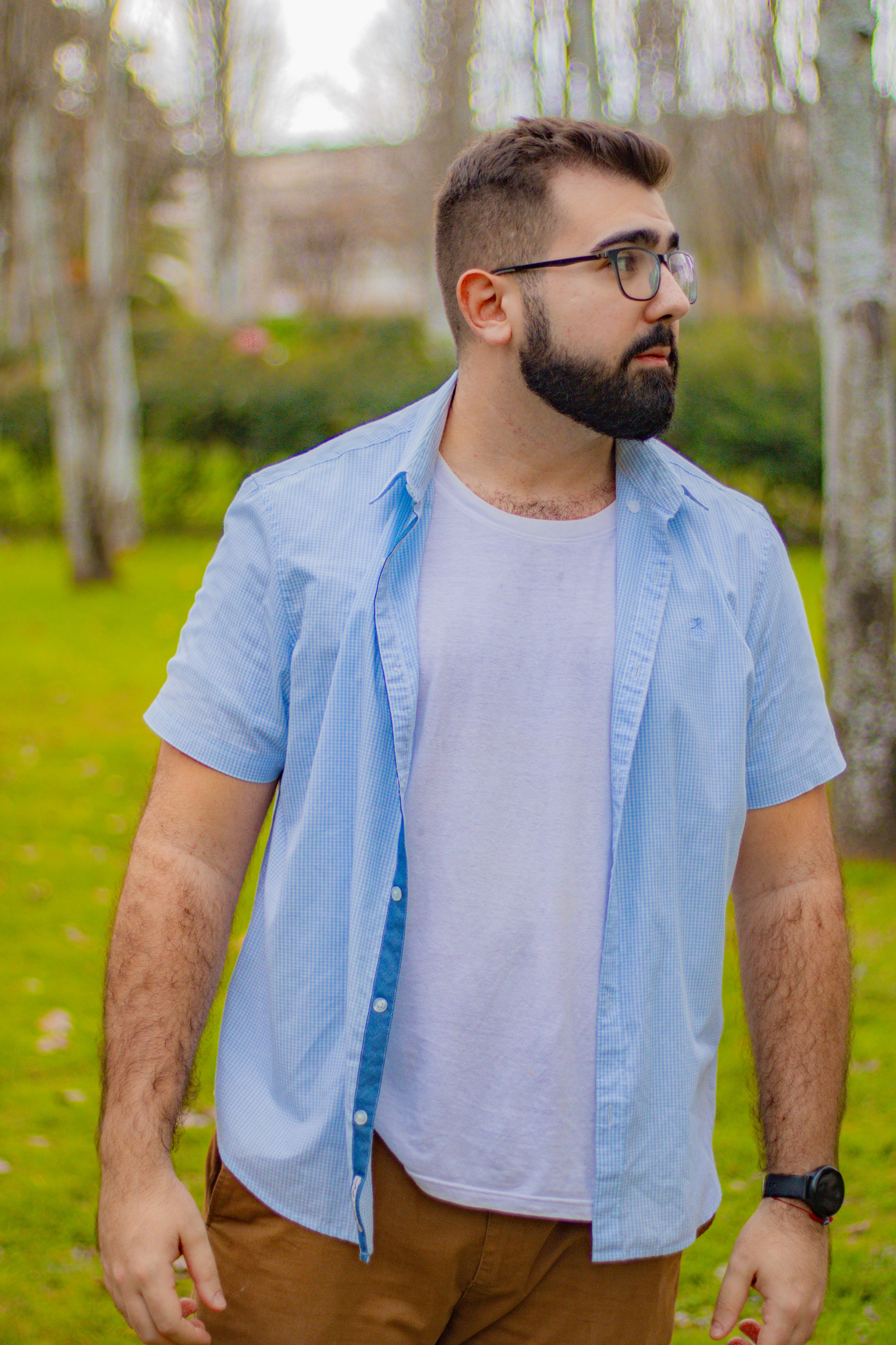 Stylish man with beard in blue shirt and glasses standing outdoors on a sunny day.