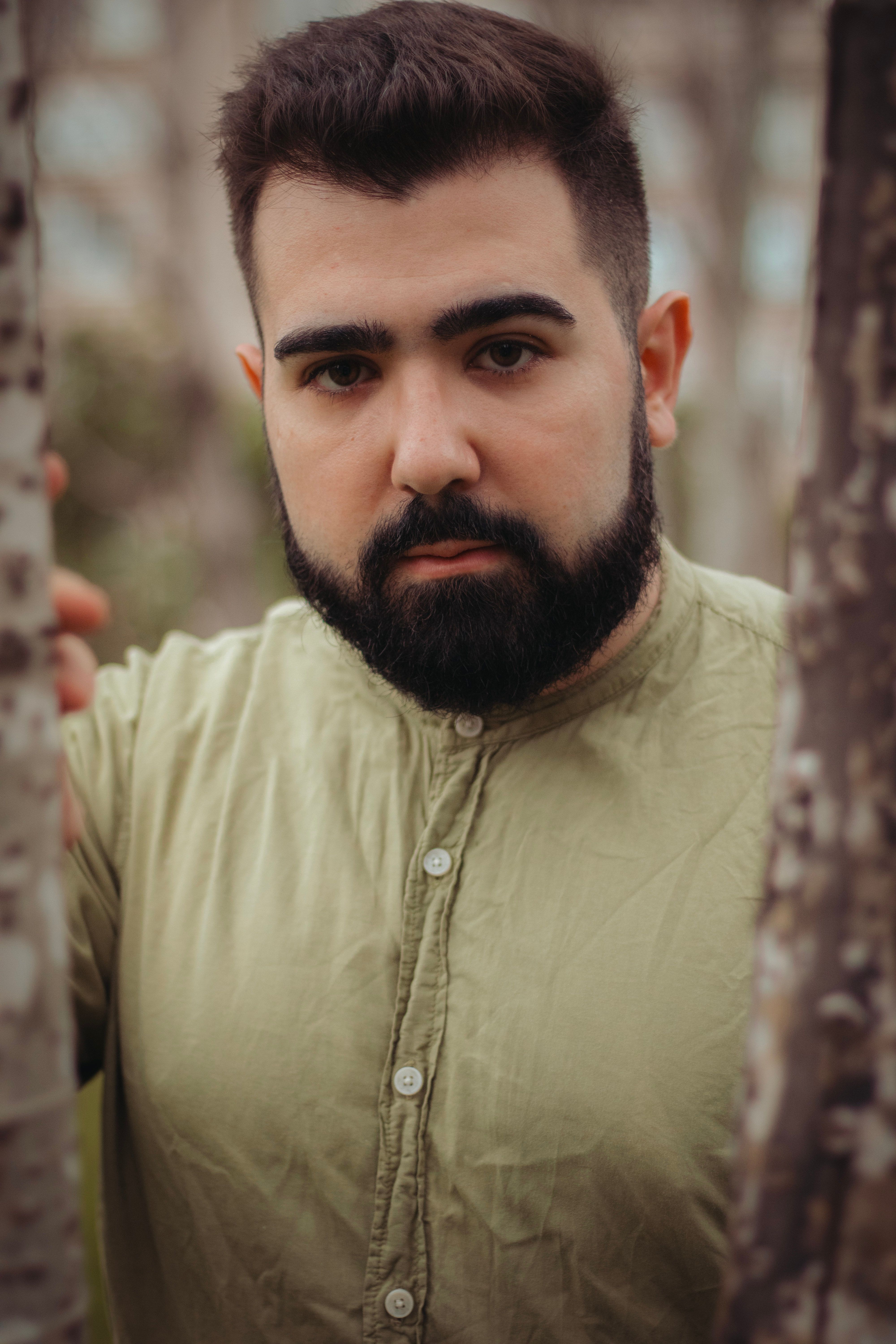 Close-up portrait of a bearded man wearing a green shirt, surrounded by trees in a natural setting.