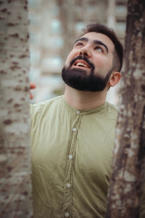 Free Portrait of a man with a beard and mustache in a green shirt, gazing upwards amidst tree trunks outdoors. Stock Photo