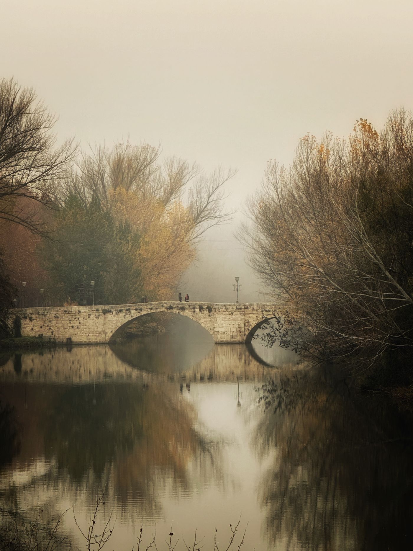 A stone bridge crossing a still river, mist across the far bank