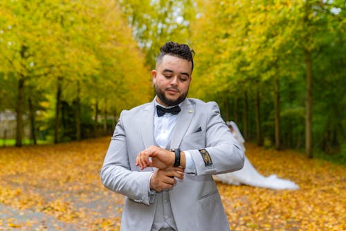 Groom Standing in a Park with Autumnal Leaves with a Bride Standing in the Background 