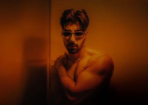 Dynamic studio portrait of a muscular man wearing sunglasses, set in warm orange lighting.