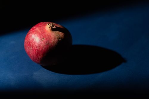 Free A red pomegranate casting a shadow on a blue tabletop under dramatic lighting. Stock Photo