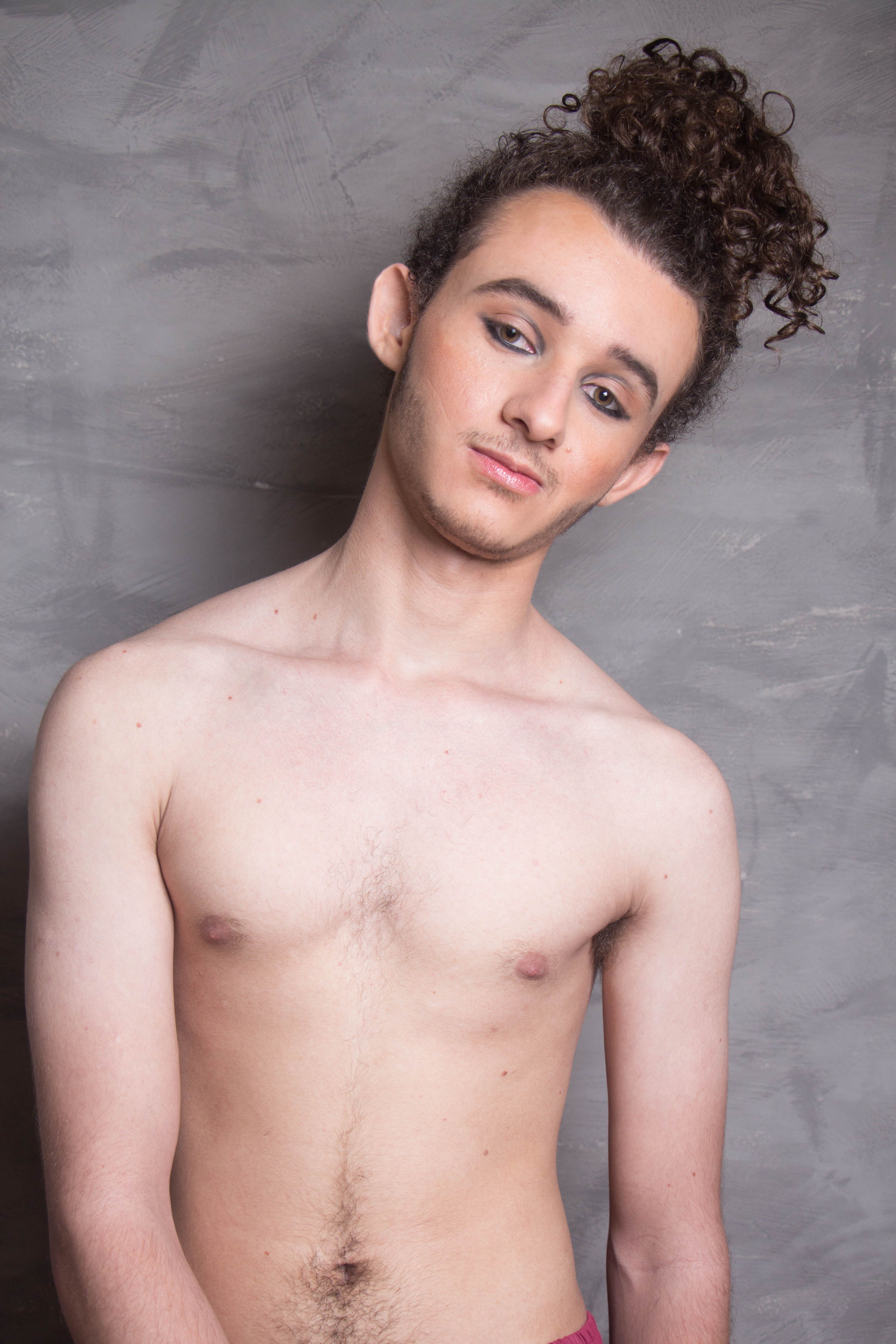 A young man with curly hair posing shirtless against a gray background.