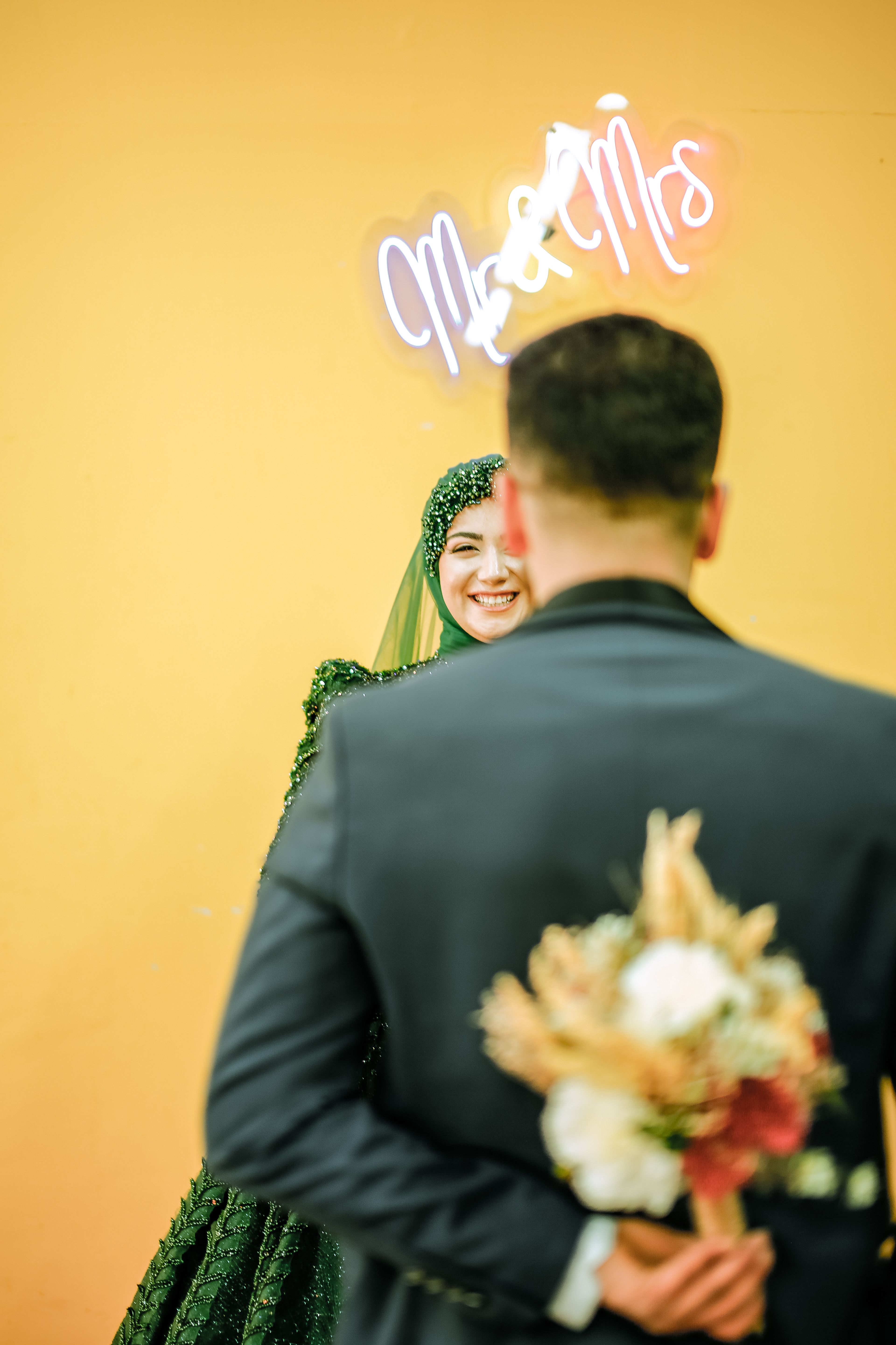 A joyful wedding scene with a bride in a green dress and hijab, smiling at her groom holding a bouquet.