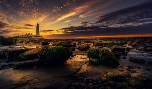 Free A stunning sunset view with a lighthouse on a rocky shore, reflecting over the water. Stock Photo