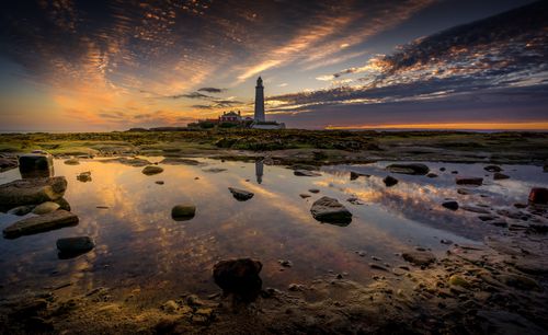 Free Beautiful lighthouse on rocky shore reflecting in water during sunset. Serene landscape with vibrant sky. Stock Photo