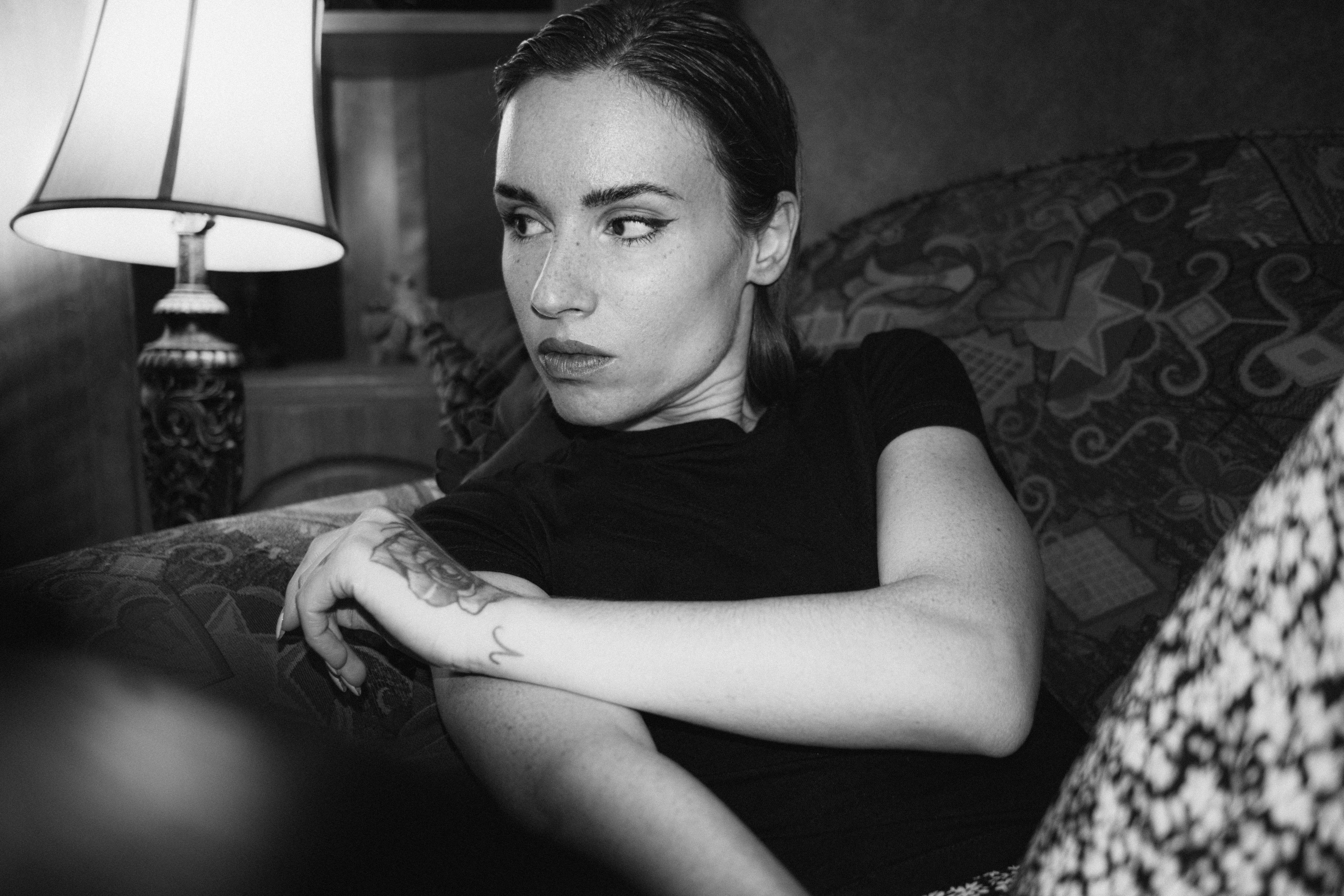 A contemplative woman with tattoos sitting on a couch in a black and white setting.