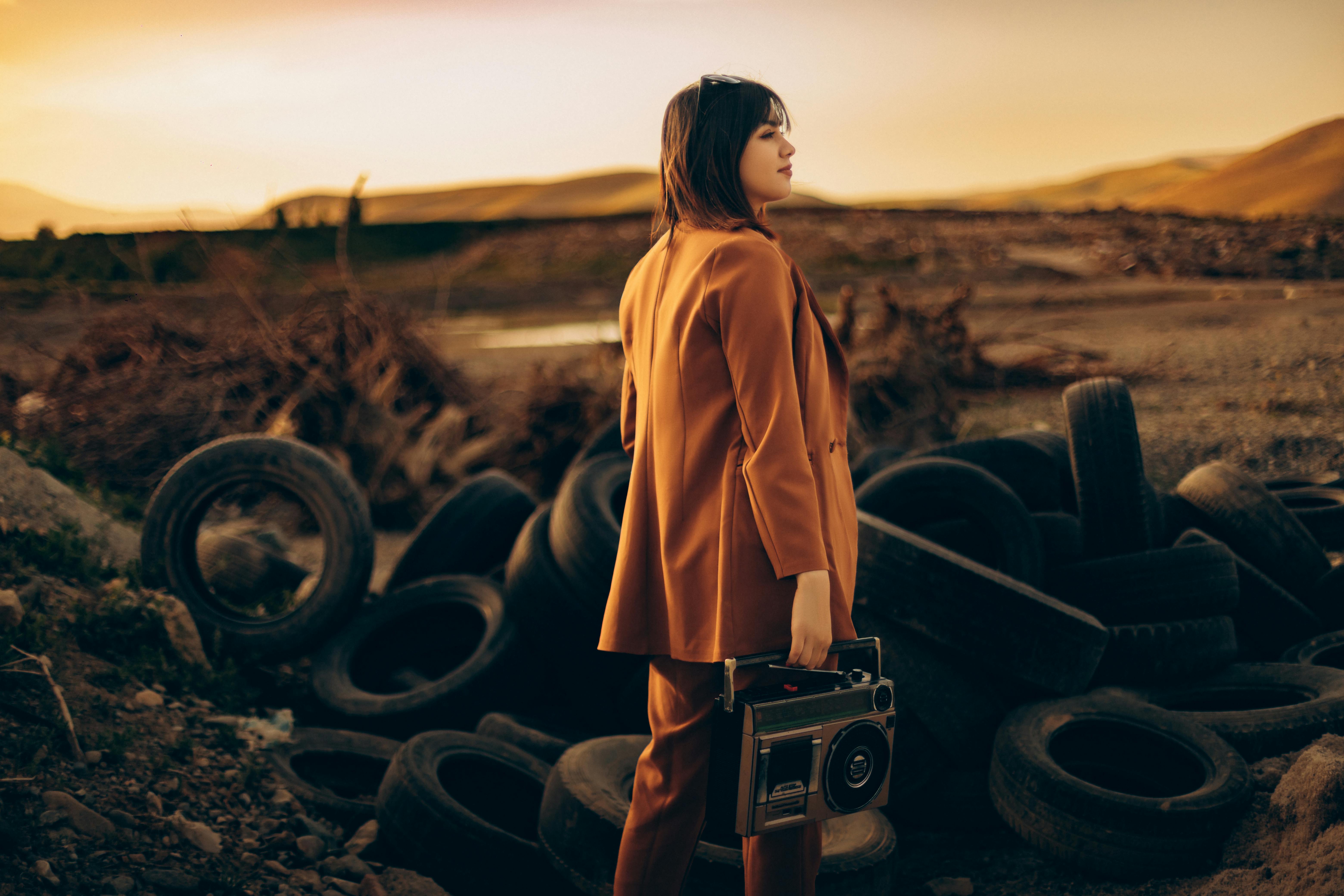 A stylish woman in an orange suit holds a vintage radio, standing near tires at sunset in Tehran.