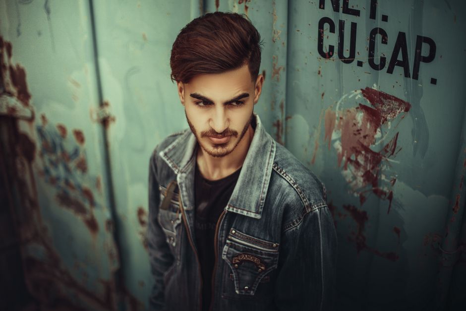 Stylish portrait of a man with brown hair wearing a jean jacket against a wall.