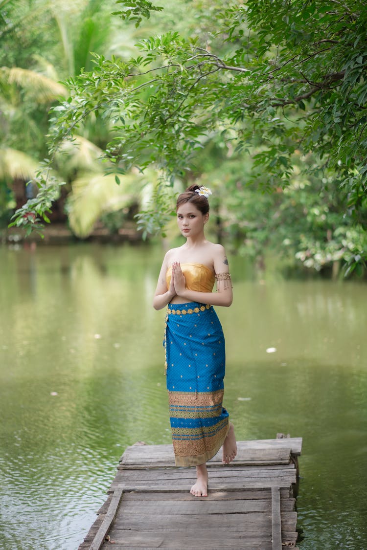 Model In A Golden Tube Top And A Blue Printed Skirt With Folded Hands Standing On One Leg On The River Pier